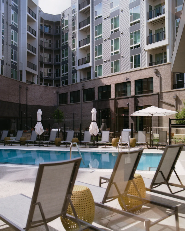 Lounge chairs and sun umbrellas surround a swimming pool in the courtyard of a large apartment building with brown brick lower stories and grayish upper stories.